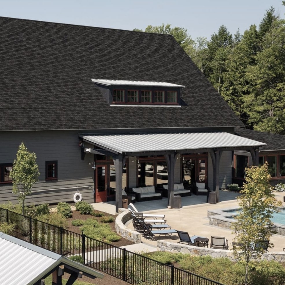 Large dark-roofed building with adjacent pool and patio area. Surrounded by trees and chairs around the pool. Fence enclosing the area.