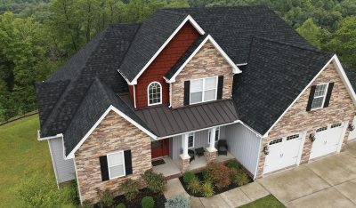 Aerial view of a large, two-story house with a brick facade, dark shingle roof, and attached three-car garage, surrounded by greenery.