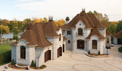 Large, elegant stone house with multiple peaked roofs, brown shingles, and arched windows set in a spacious driveway. Tree-lined background featuring autumn foliage.