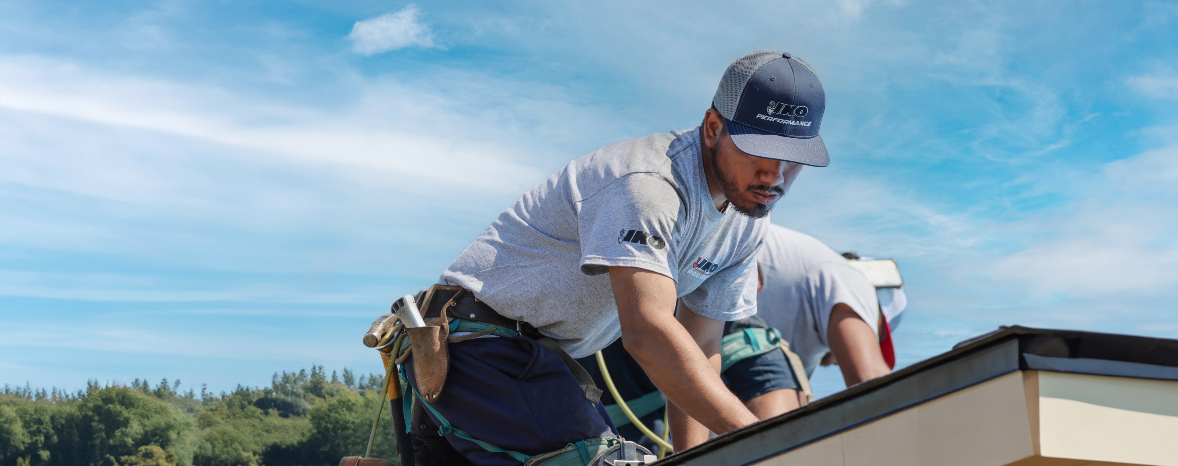 Two workers in gray shirts and caps are focused on roofing solutions under a clear blue sky. One is kneeling near a roof's edge, with trees visible in the background.