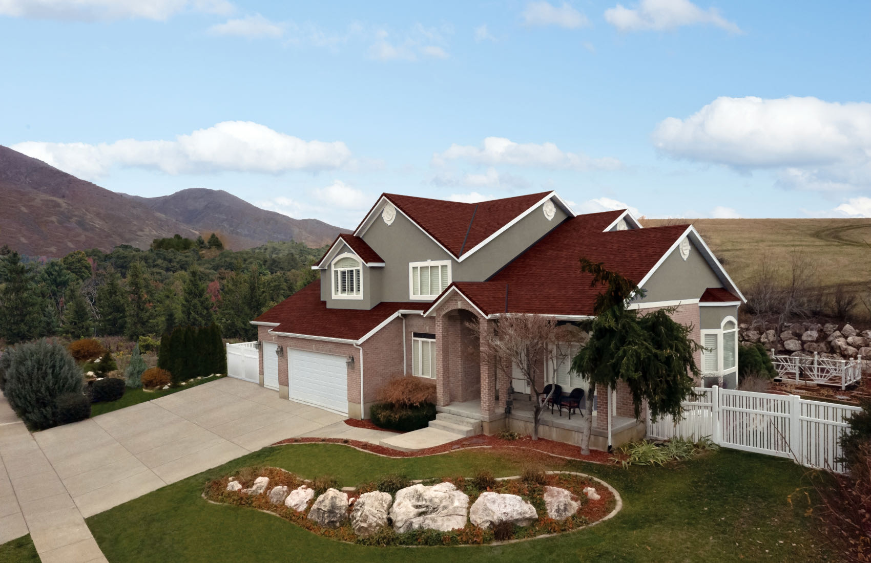 Large suburban home with a red roof, white trim, and attached garage. The front yard features a rock garden like an outdoor gallery. Mountains and a cloudy sky serve as the breathtaking backdrop.