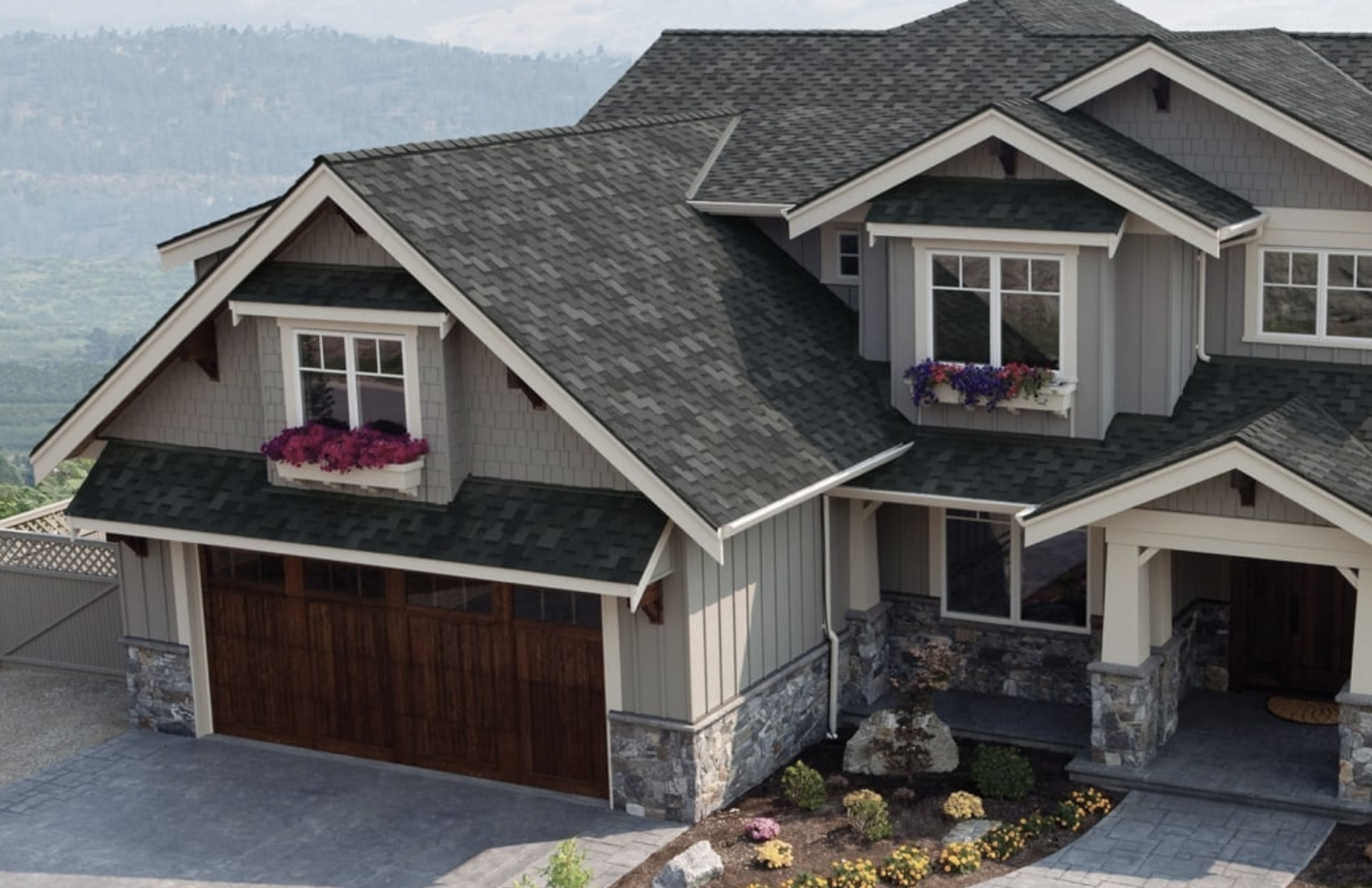 A two-story house with a gray roof and stone accents showcases wooden garage doors in a distinct style. Board trim enhances the exterior’s charm, while the front yard features small shrubs and flowers in bloom.