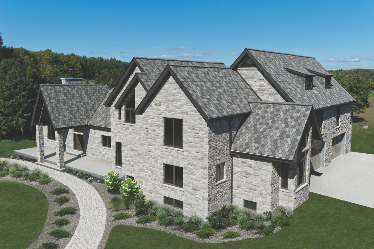 A large stone house with multiple peaked roofs and dormers, crowned with elegant slate tiles, is surrounded by a paved walkway and lush green lawn.