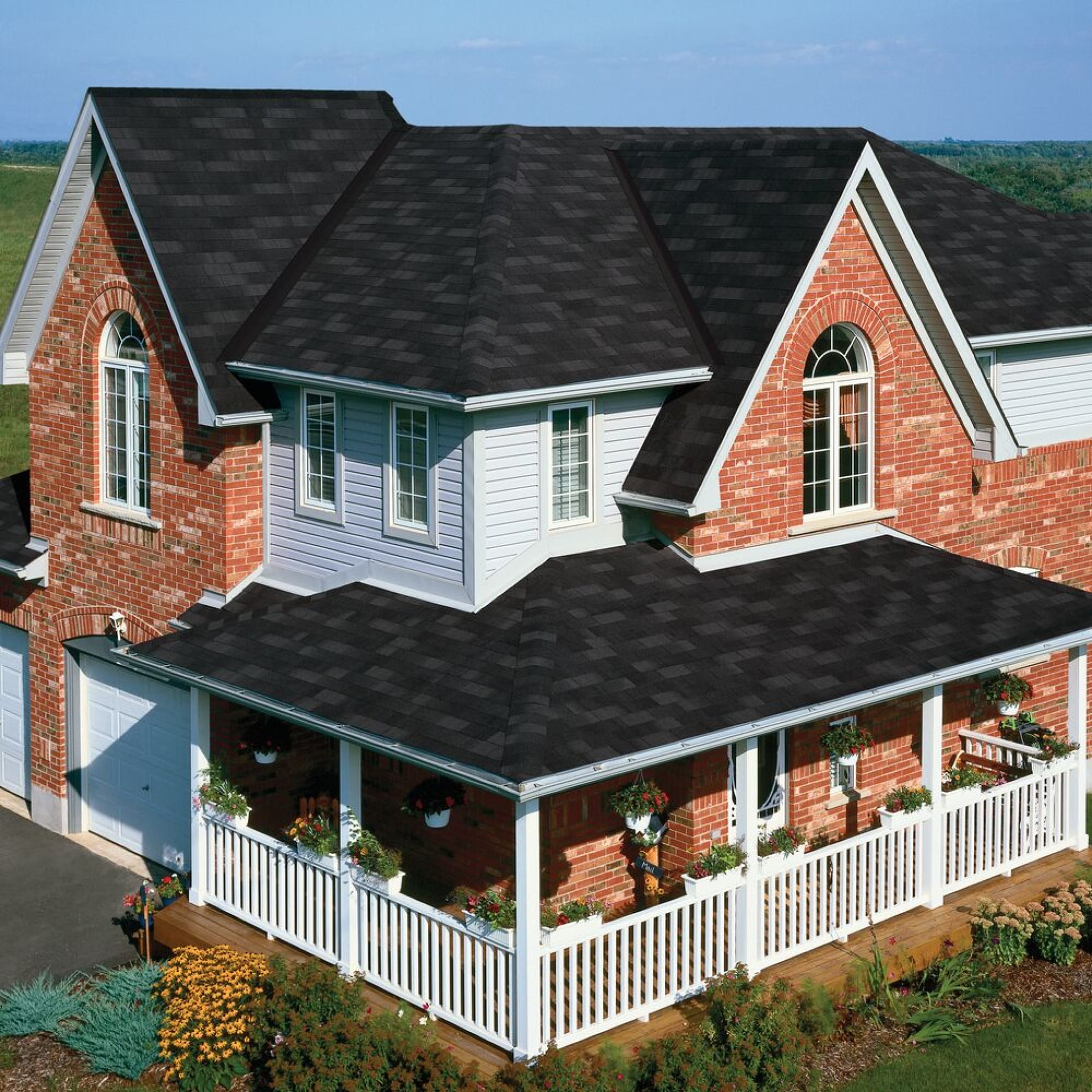 A two-story red brick house with white trim and a Dual Black asphalt shingle roof is surrounded by a charming white picket fence. The front porch showcases hanging flower baskets, while Marathon Plus AR greenery adds to the serene atmosphere.