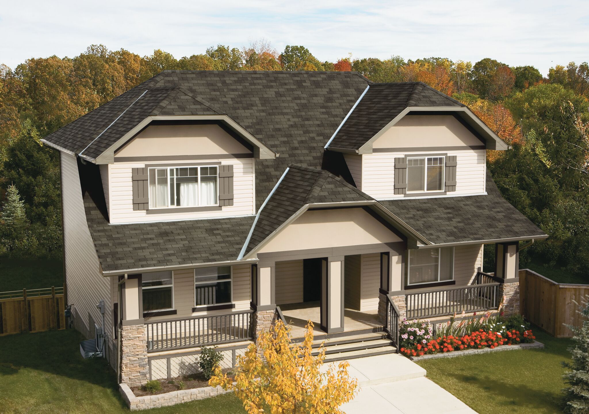 The charming two-story house features a grey Weatherwood roof and white walls with stone accents. A front porch with wooden railings, surrounded by trees and flowers, invites you in. Steps lead to the welcoming front door for the perfect blend of nature and comfort.