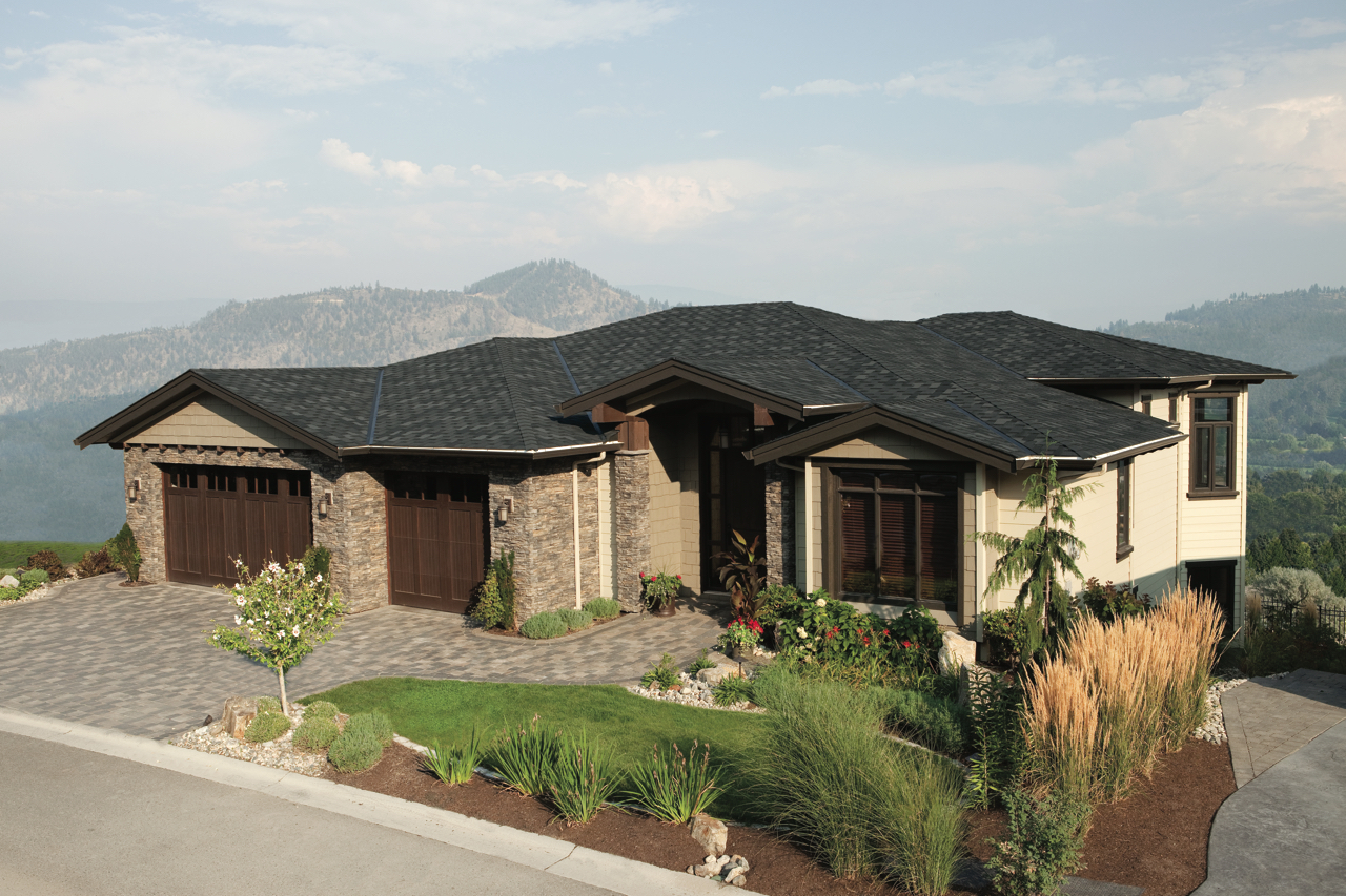 A modern single-story house with a stone facade, dark roof, and three-car garage, surrounded by a landscaped yard. Mountains and greenery are visible in the background.