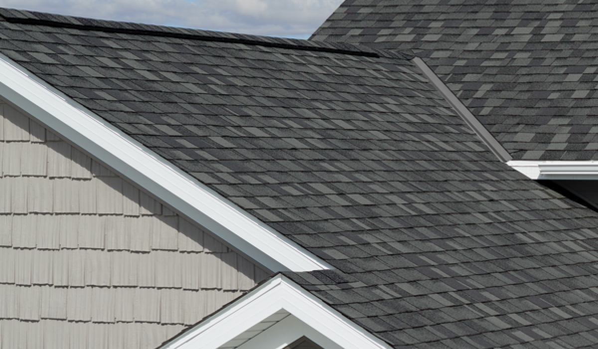 Close-up of a house roof with dark gray asphalt shingles, contrasting with light gray wooden siding.