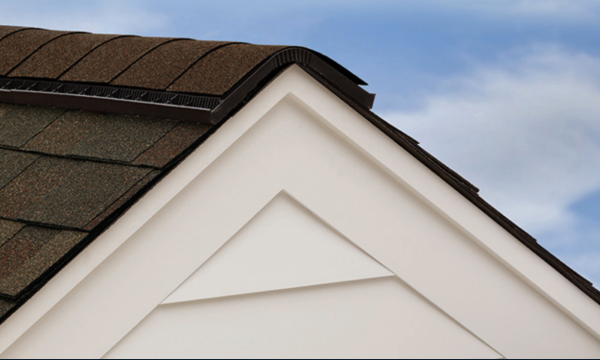 Close-up of a house roof corner showcasing Hip and Ridge™ asphalt shingles in brown, with beige siding underneath against a clear blue sky.