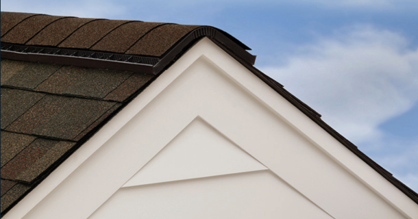 Close-up of a house roof corner showcasing Hip and Ridge™ asphalt shingles in brown, with beige siding underneath against a clear blue sky.