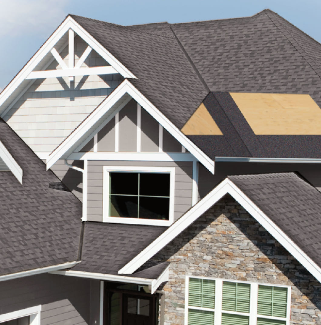 A house with a gabled roof, grey shingles, stone facade, and green window shutters offers robust storm shield features. One section of the roof is currently under construction, ensuring enhanced weather protection.