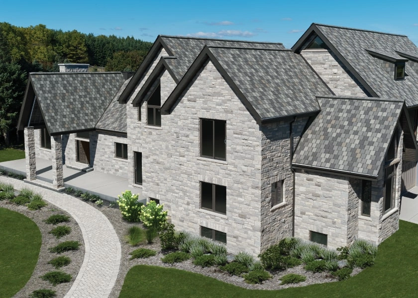 A large stone house with multiple gabled roofs featuring Crowne Slate tiles and a paved walkway, surrounded by manicured lawns and trees under a clear blue sky.