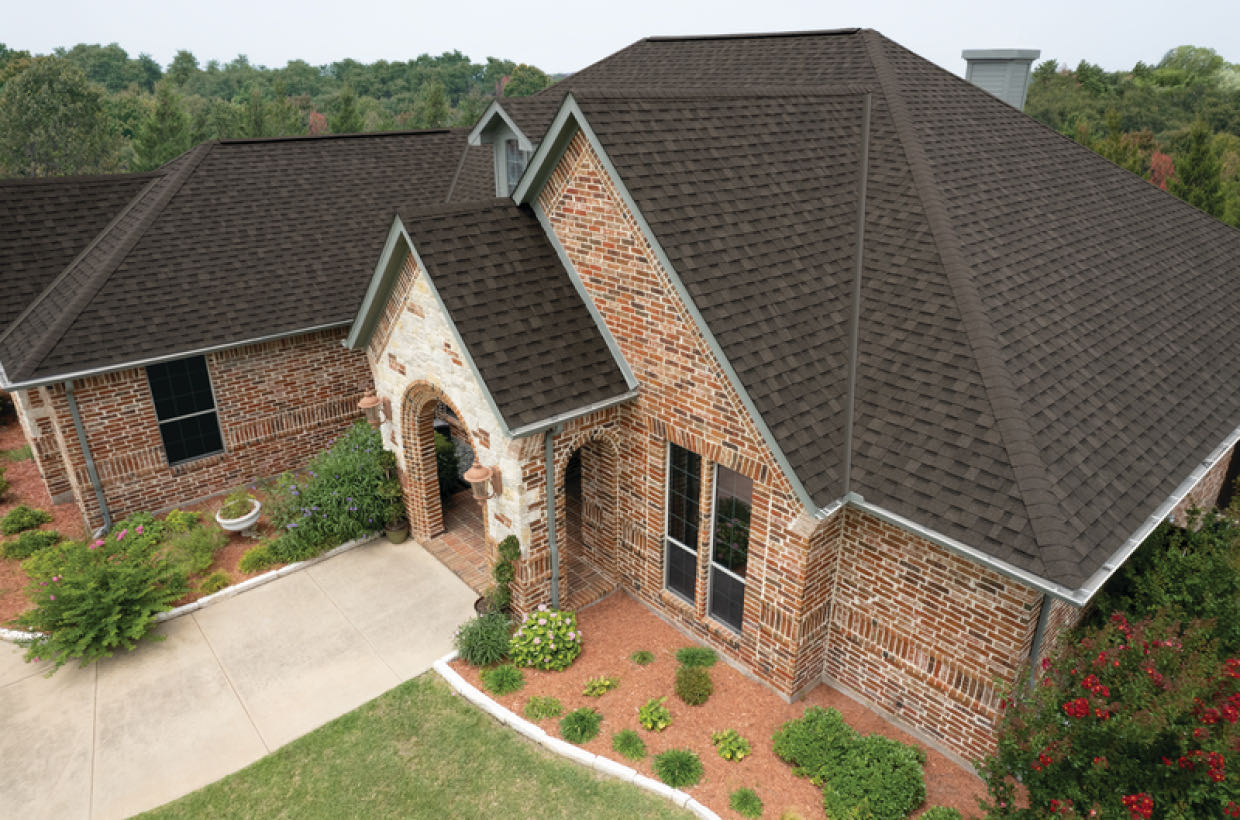 Brick house with a large taupe gabled roof, surrounded by shrubs and a small driveway, set against a backdrop of trees.