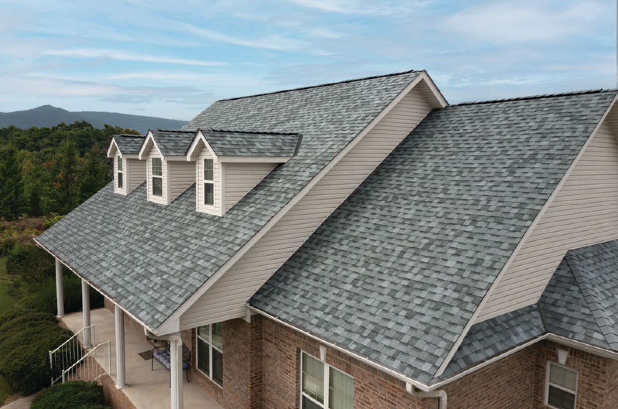 A house with a steep gray shingle roof, featuring three dormer windows. Brick exterior walls and a porch are visible. Trees and mountains are in the background under a cloudy sky.