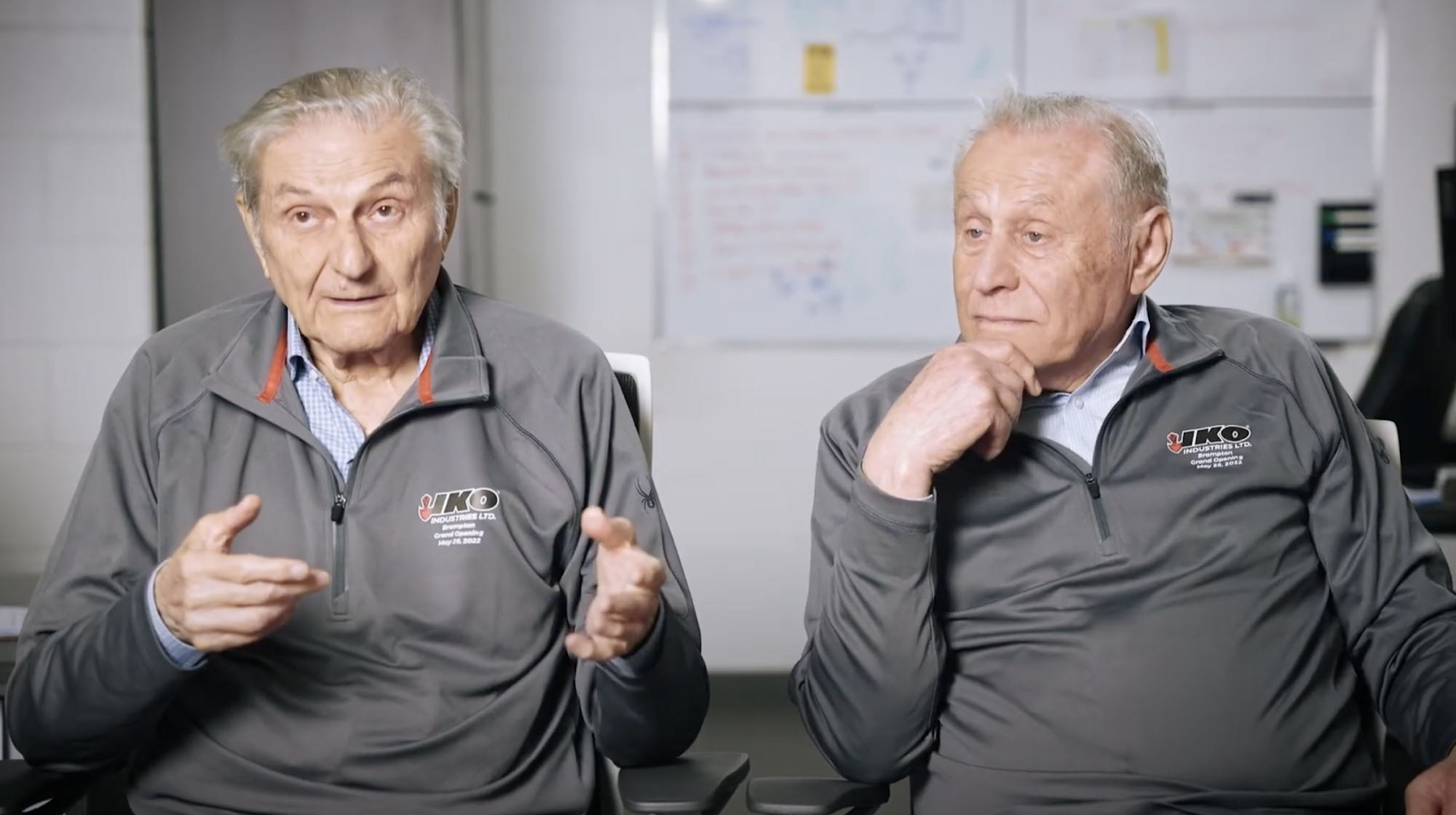 In an office setting with a whiteboard in the background, two elderly men in matching gray shirts sit side by side. One gestures animatedly while discussing IKO, as the other listens intently, hand to chin, contemplating the difference in their viewpoints.