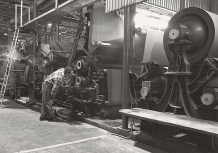 A person operates large industrial machinery in a factory setting, with gears and rollers visible. A ladder leans against the equipment, highlighting the precision needed to make a difference in sectors like roofing production with IKO's advanced technology.