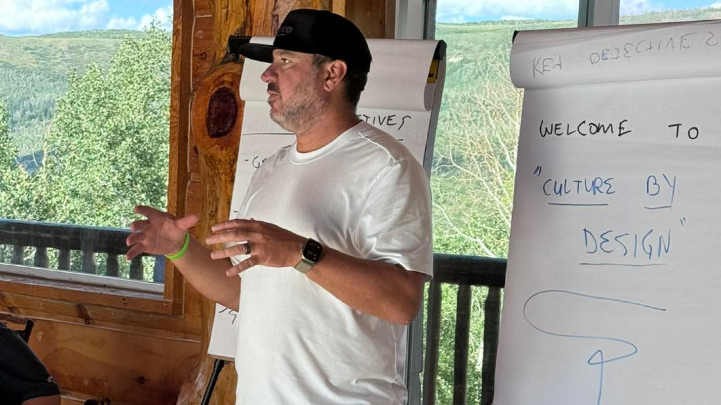 A man in a white T-shirt speaks passionately about "Culture by Design" in front of flip charts, with large windows framing trees and hills. His insights reflect the core values shared by ROOFPRO, emphasizing how a strong culture can amplify roof benefits for both team and client success.