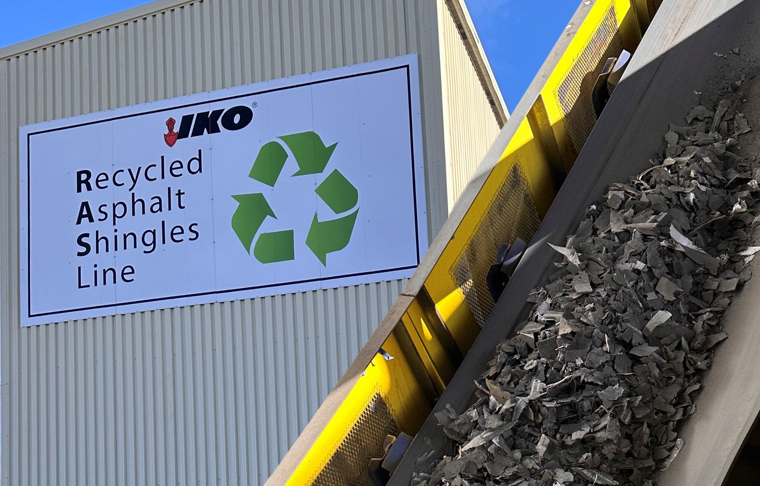 A conveyor belt moves recycled asphalt shingles at a facility. A sign reads "IKO Recycled Asphalt Shingles Line" with a recycling symbol.