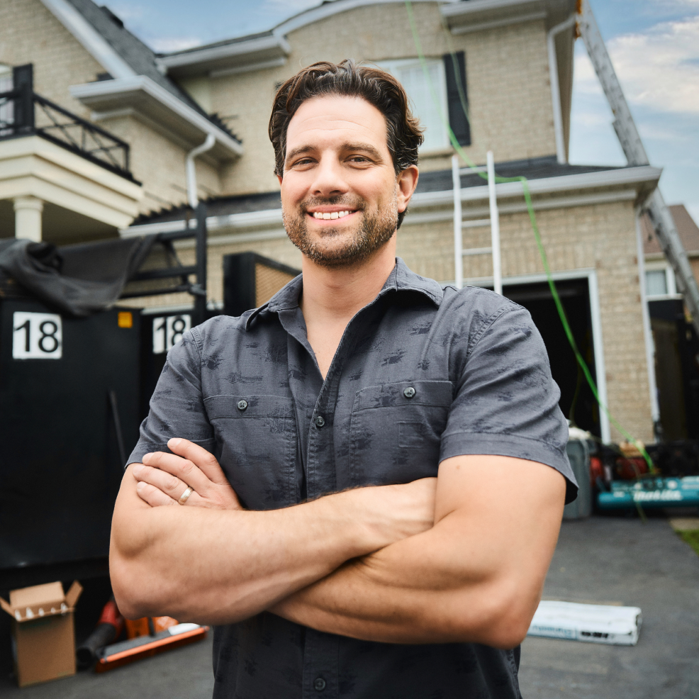 A man in a gray shirt stands with arms crossed, smiling confidently like Scott McGillivray in front of a house under construction.