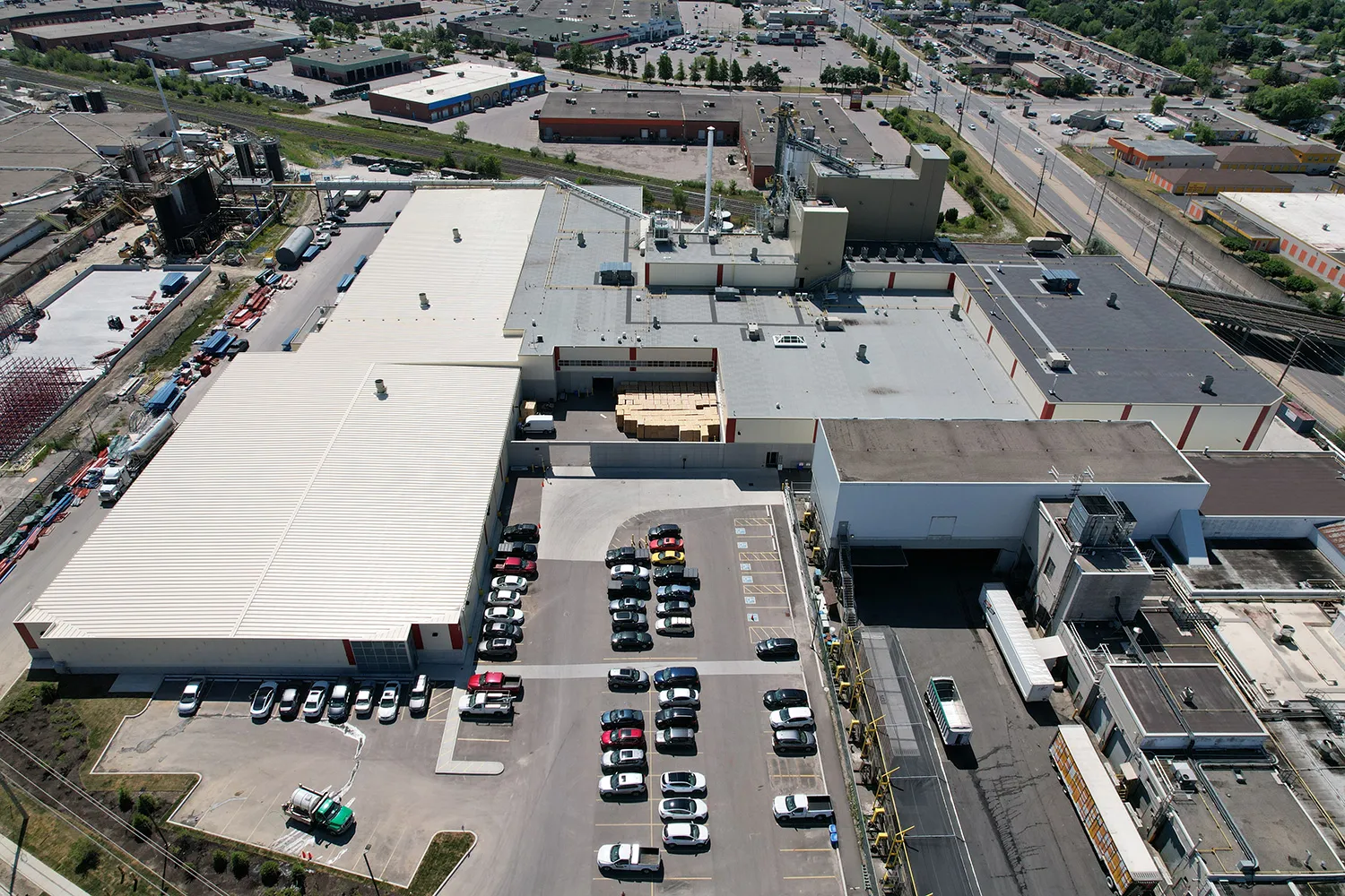 Aerial view of an industrial facility with multiple buildings, parking areas filled with cars, and surrounding roads in an urban setting.