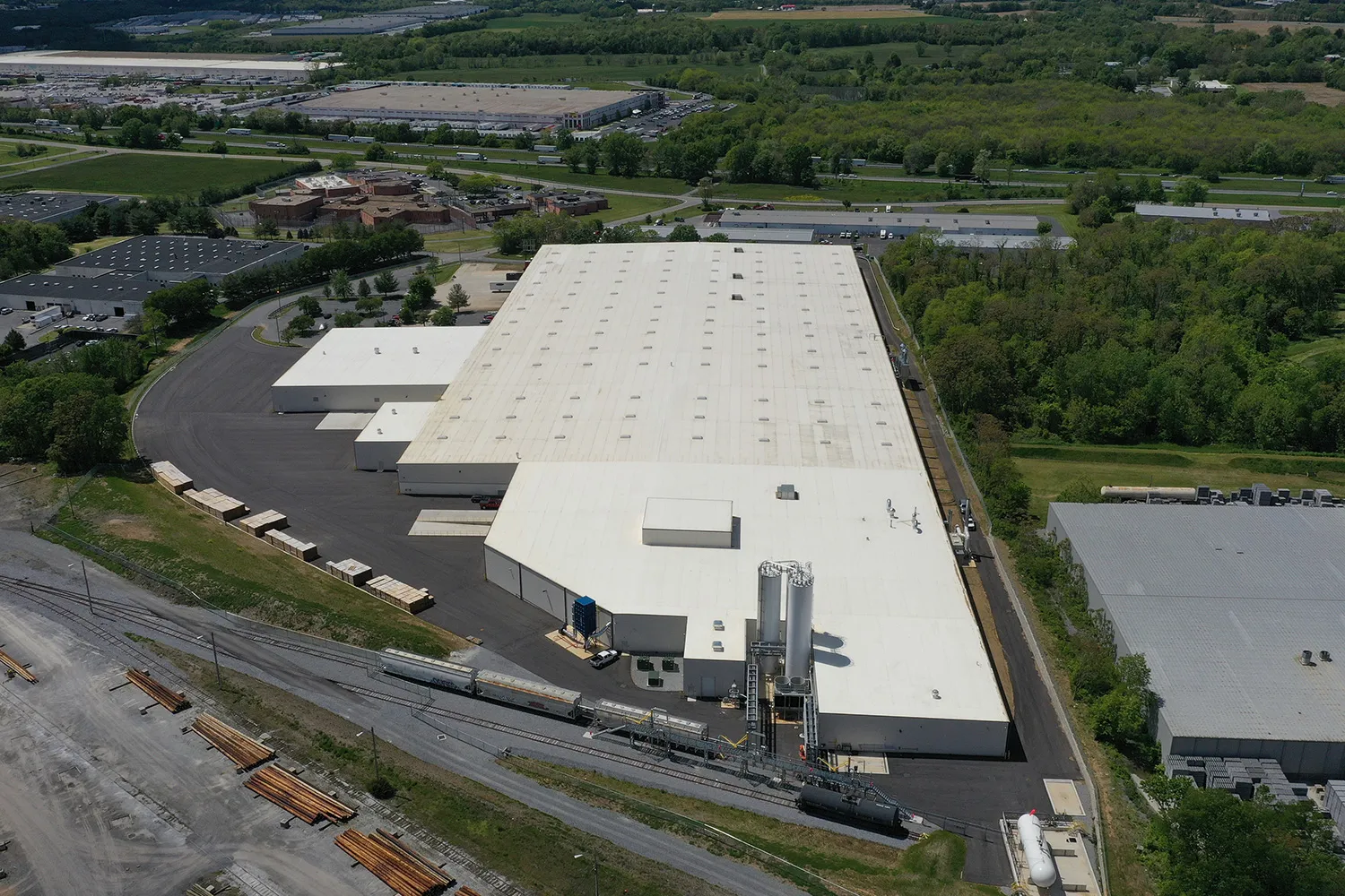 Aerial view of a large industrial warehouse with a white roof, surrounded by roads and greenery, with trains and construction materials nearby.