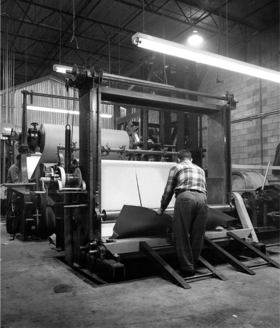 A worker in a plaid shirt operates a large industrial paper cutting machine in a factory setting.