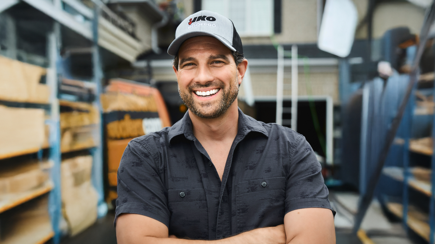 A smiling man in a dark shirt and cap stands outdoors, arms crossed with inspiration, in front of industrial equipment.