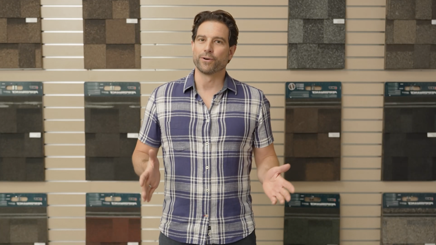 A man in a plaid shirt stands in front of a ROOFPRO wall display, highlighting the benefits of various roofing shingles.