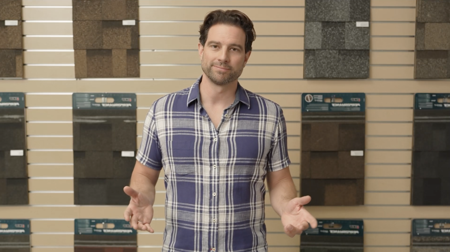 A man in a plaid shirt stands in front of a display of various roofing shingles, gesturing with his hands.