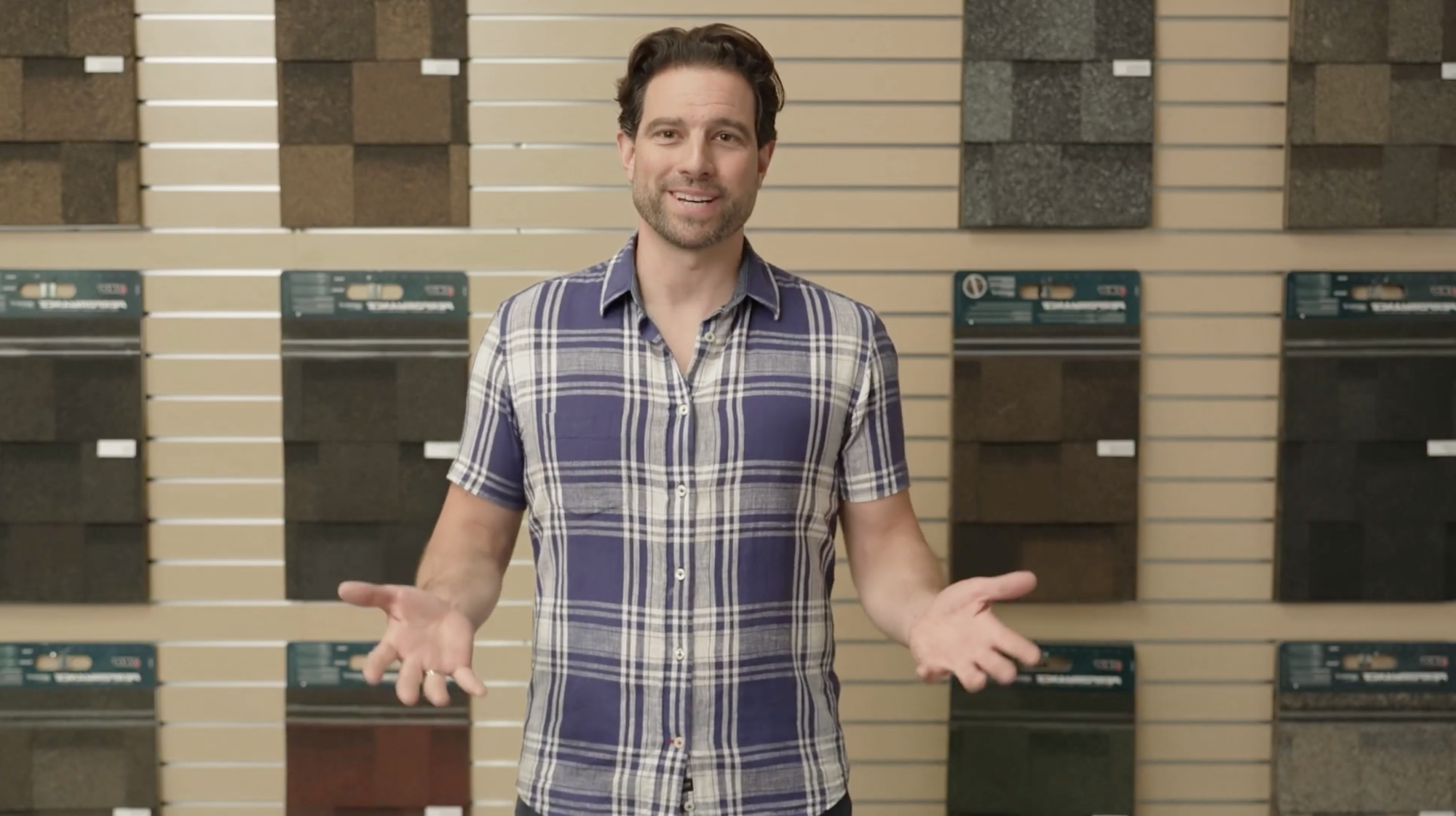 A person stands in front of a wall display of roofing materials, wearing a blue and white plaid shirt, gesturing with open hands like real estate expert Scott McGillivray discussing home renovation tips.