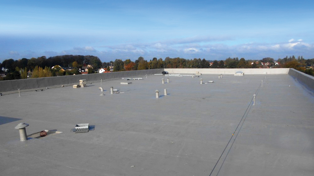 Flat rooftop view with various vents and pipes; a clear sky and distant treetops and houses in the background.