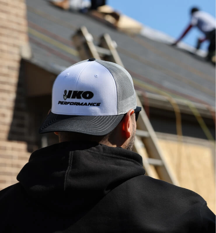 Person in cap observing roofing work on a house, with ladders and workers visible in the background.