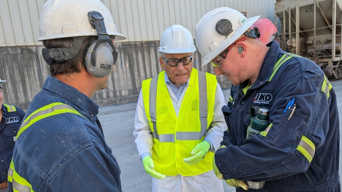 Three workers in safety gear and helmets stand talking outside an industrial setting, exploring career opportunities. One of them wears a bright yellow vest.