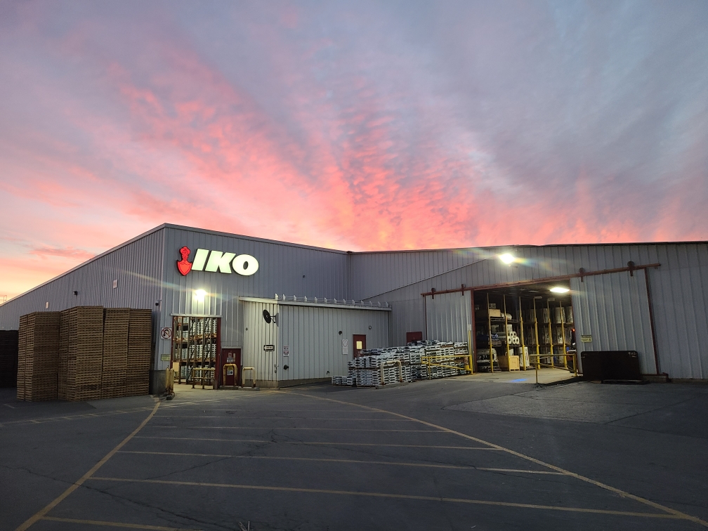 The exterior of a large industrial warehouse, proudly displaying the "IKO" sign, stands amidst stacks of materials. A vibrant pink and blue sunset paints the backdrop, highlighting IKO's commitment to sustainable construction as advocated by the Canada Green Building Council.