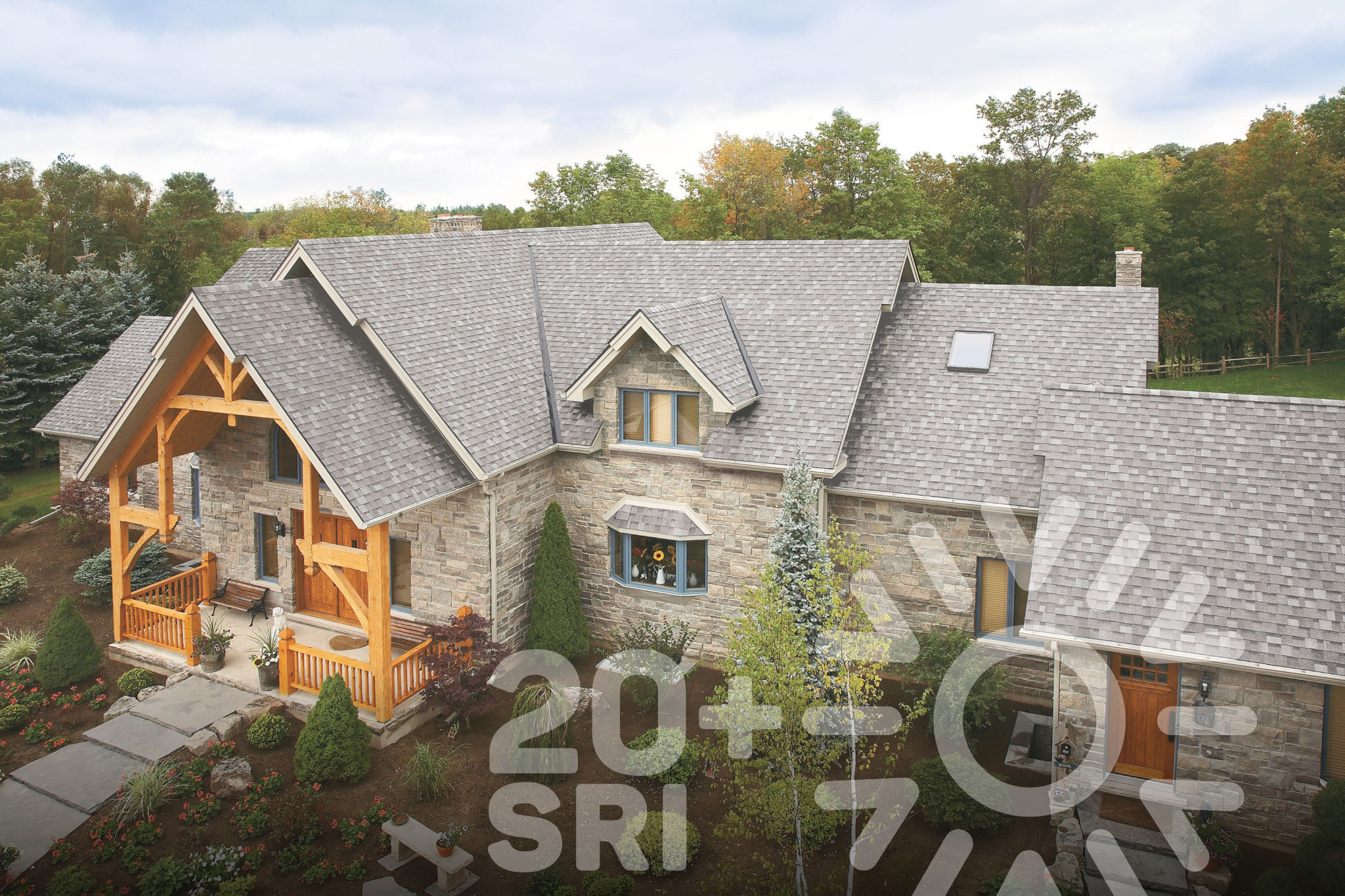 Aerial view of a large stone house with a gray Shingle Line roof, wooden porch details, surrounded by trees. Text overlay reads "20+ SRI" with a sun symbol, highlighting how New Dynasty Cool Colors elevate roofing aesthetics.