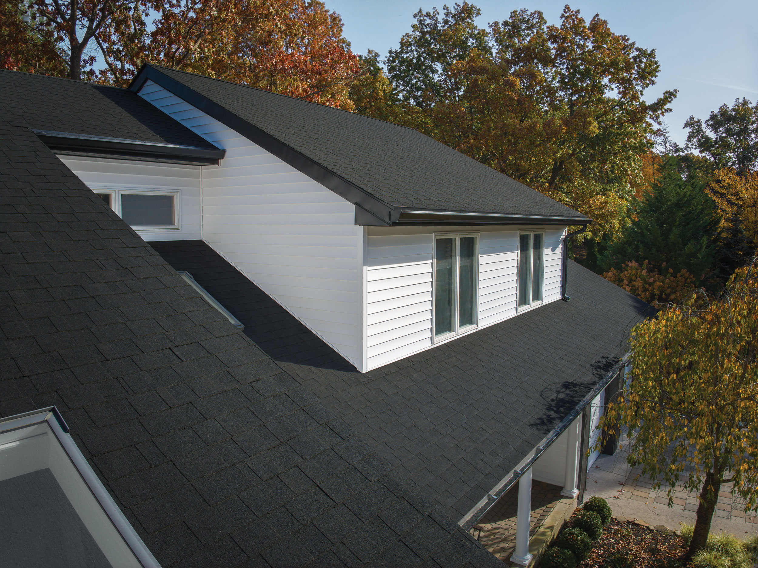 House with Dynasty Shingles, white siding, and large windows. Surrounded by autumn trees. The prominent roof design in the foreground features a matte black finish.