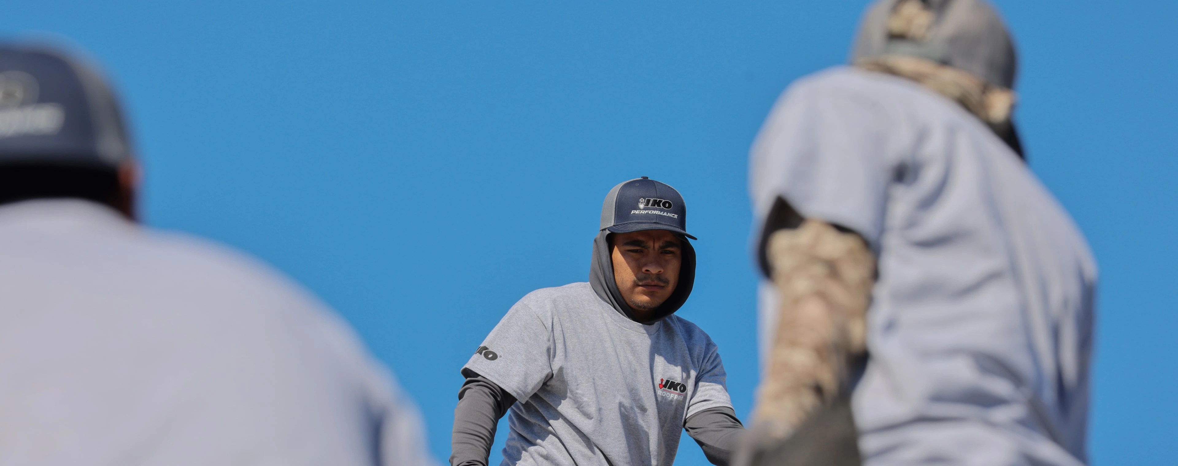 A construction worker in a gray shirt and cap stands outdoors under a clear blue sky, framed by blurred figures in similar attire.