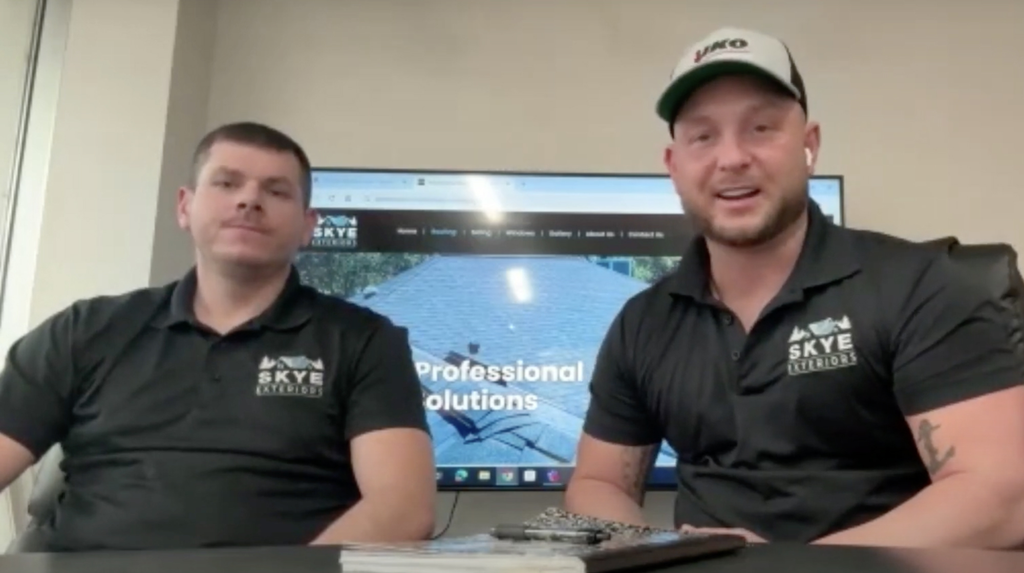 Two professionals in black Skye Exteriors shirts sit at a table, with a computer displaying an IKO roofing website in the background.