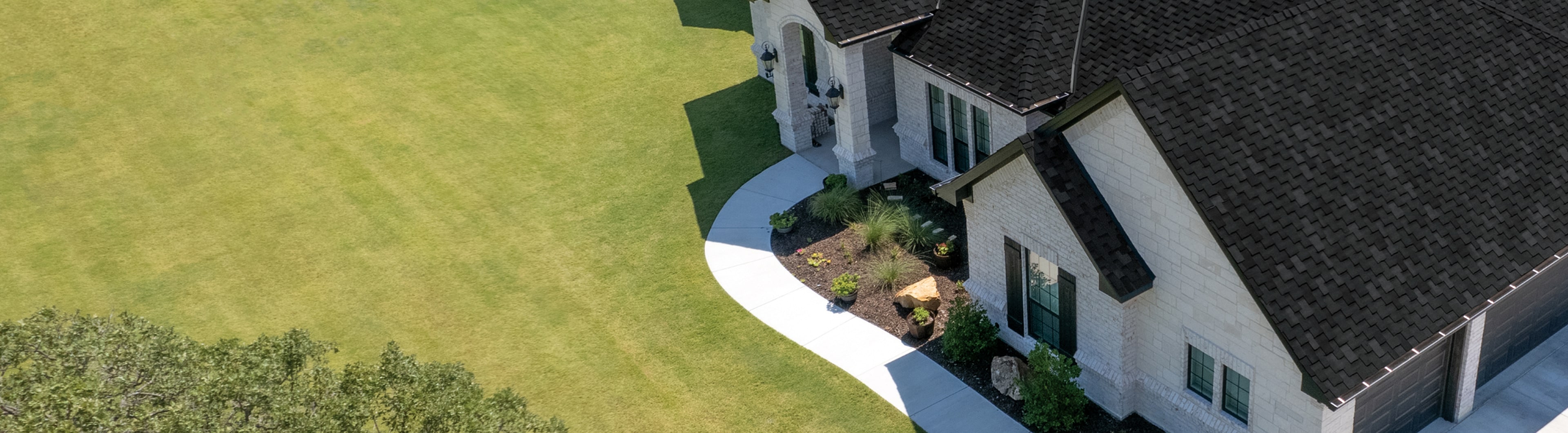 Aerial view of a home with a dark roof, light exterior, and green lawn. The yard features a curved concrete pathway leading to the entrance, resembling an outdoor gallery with small shrubs and plants near the house.