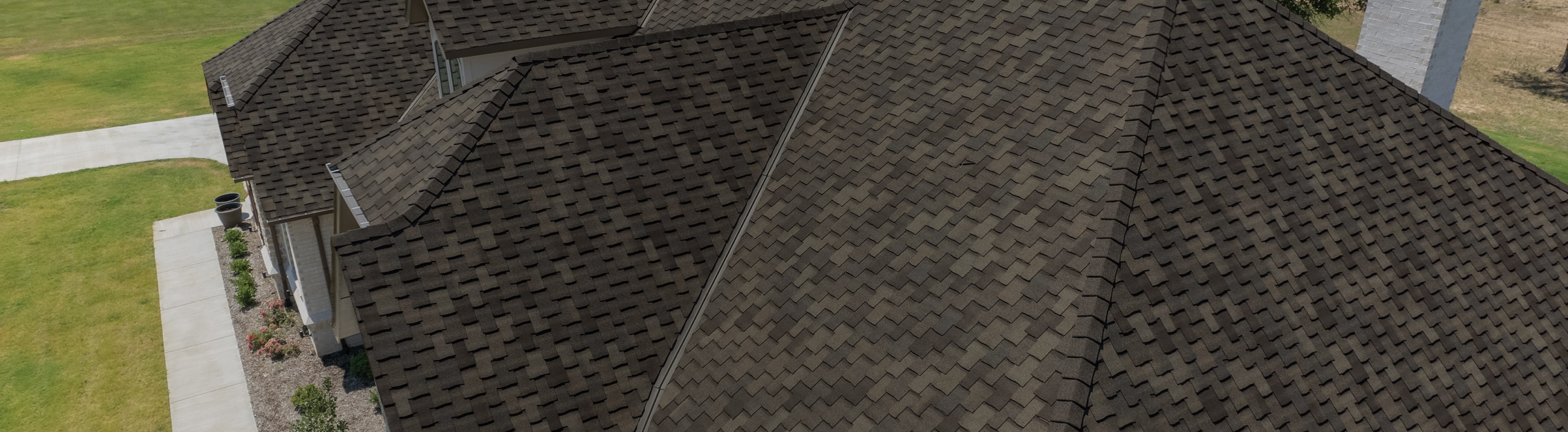 Aerial view of a dark-colored shingled roof on a residential house, beautifully contrasting with the vibrant green lawn and concrete driveway, creating a stunning gallery of natural and architectural color harmony.