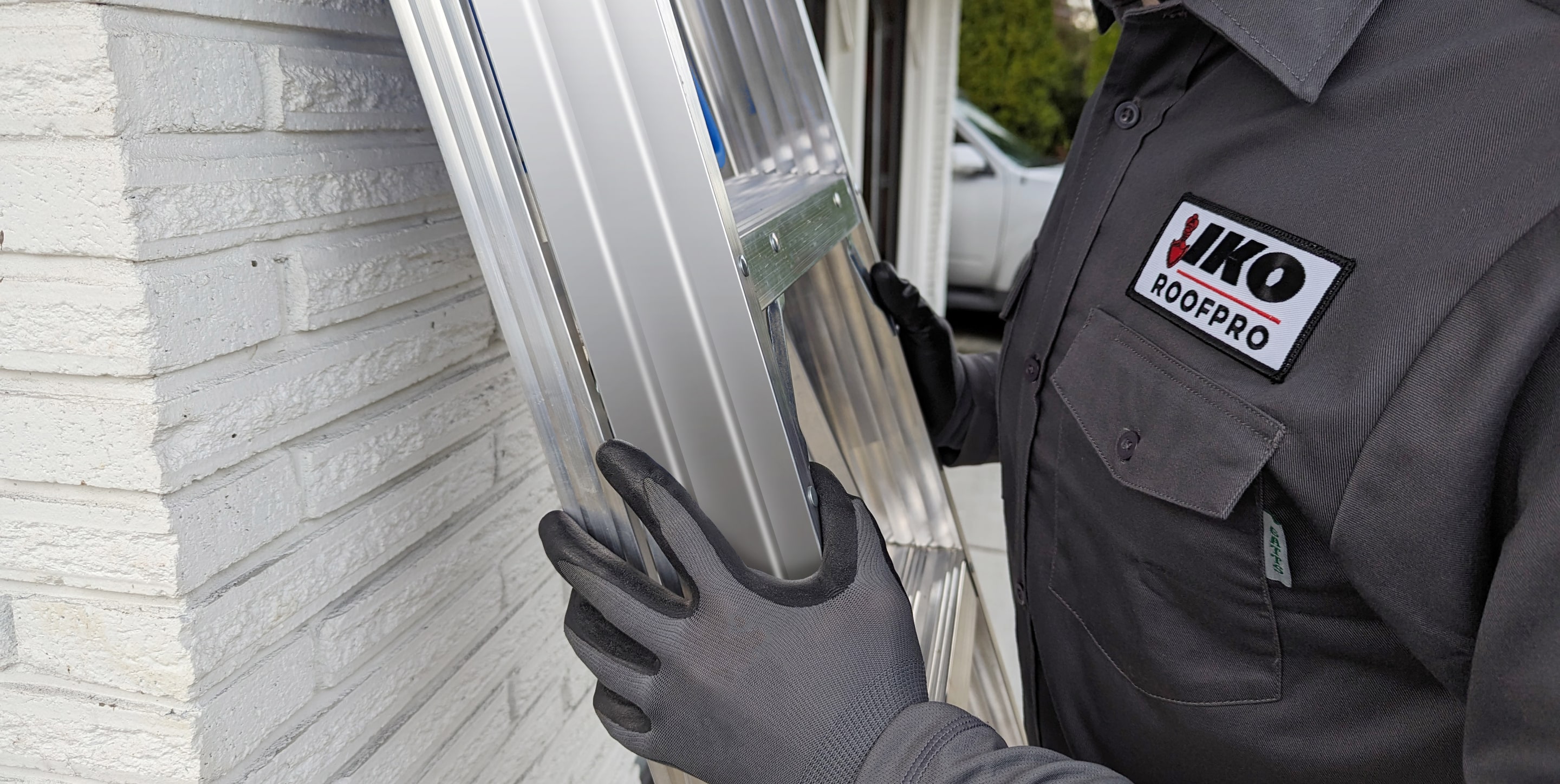 A person wearing gloves holds an aluminum ladder against a white brick wall. The individual's shirt has a