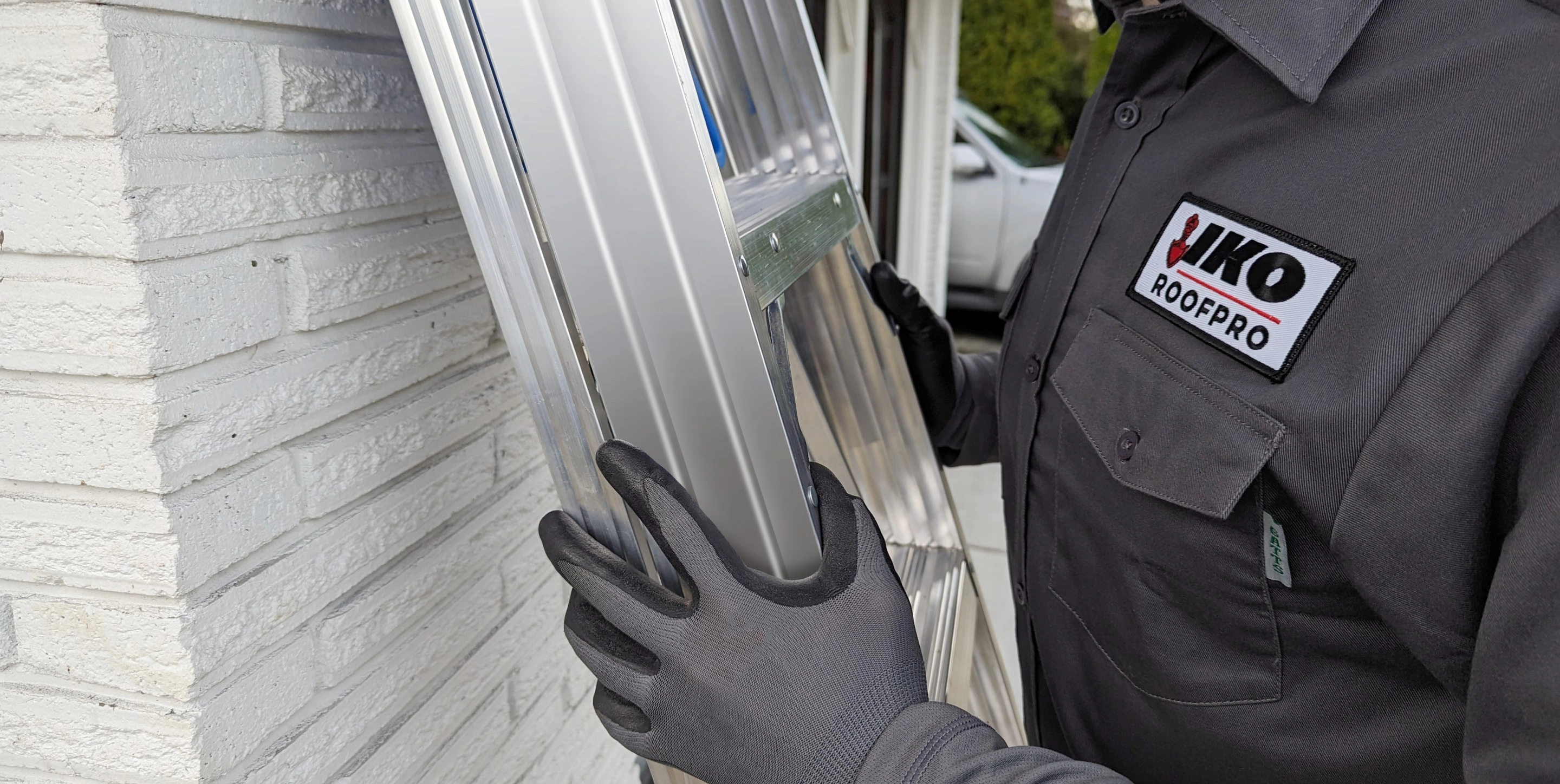 A person wearing gloves holds an aluminum ladder against a white brick wall. The individual's shirt has a 