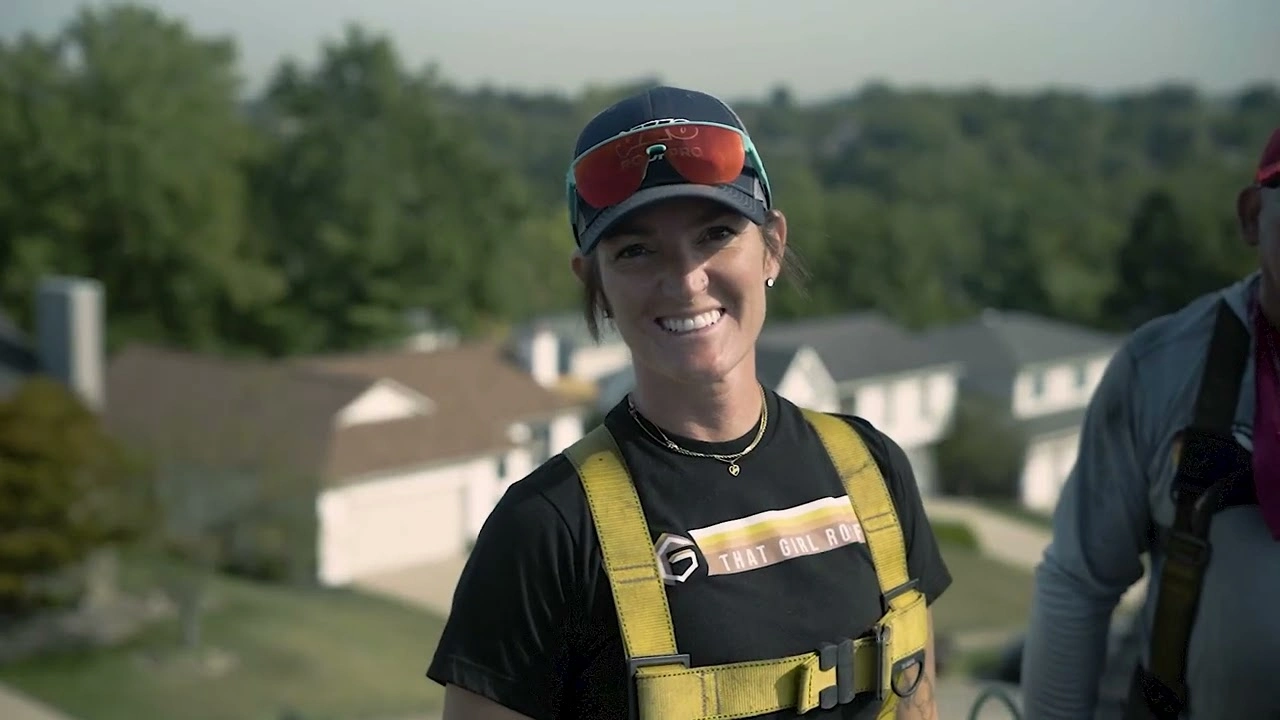 A person in a safety harness and sunglasses smiles confidently on a rooftop, showcasing the precision of That Girl Roofing, with houses and trees elegantly spread out in the background.