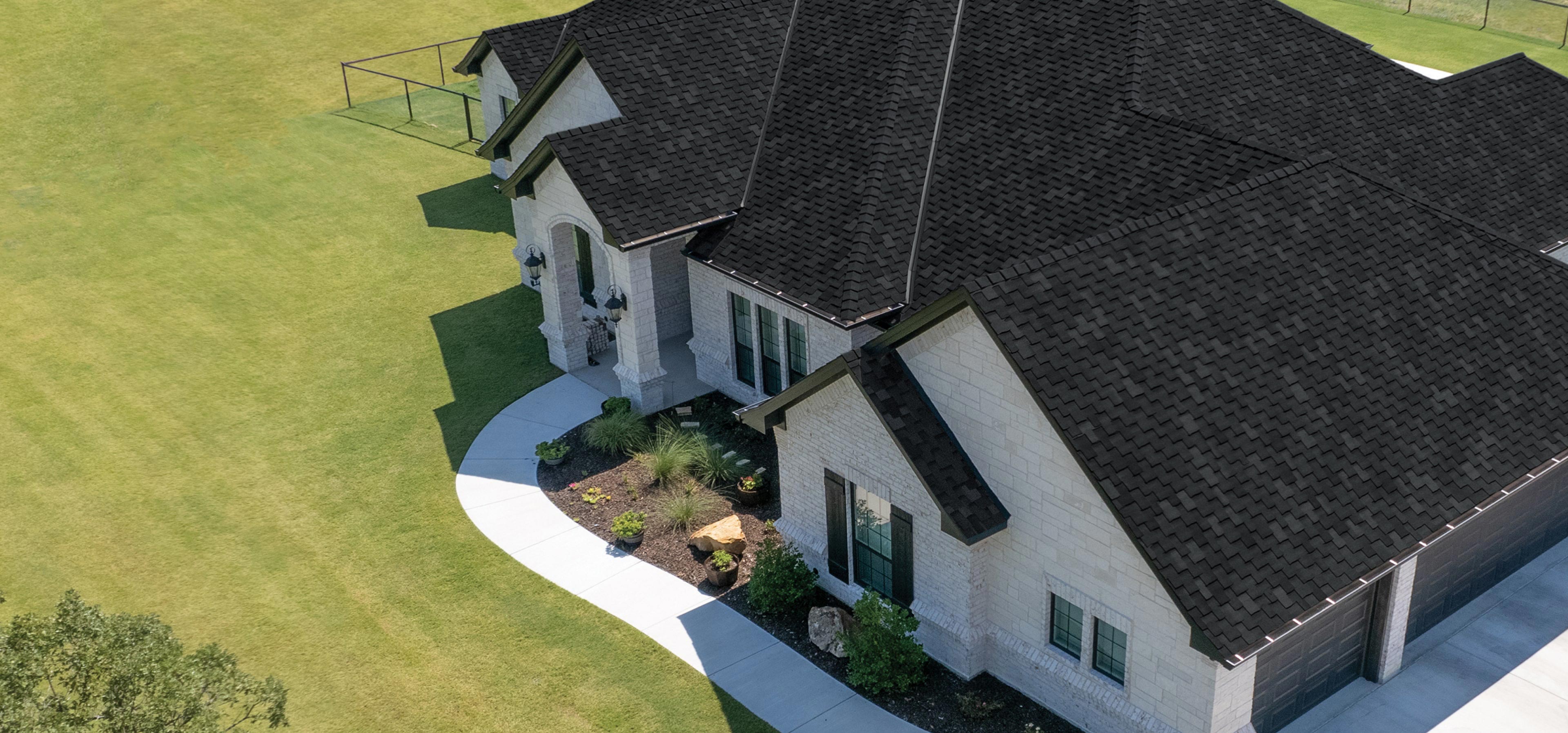 Aerial view of a large, single-story house with a dark shingled roof, light brick exterior, and surrounding green lawn.